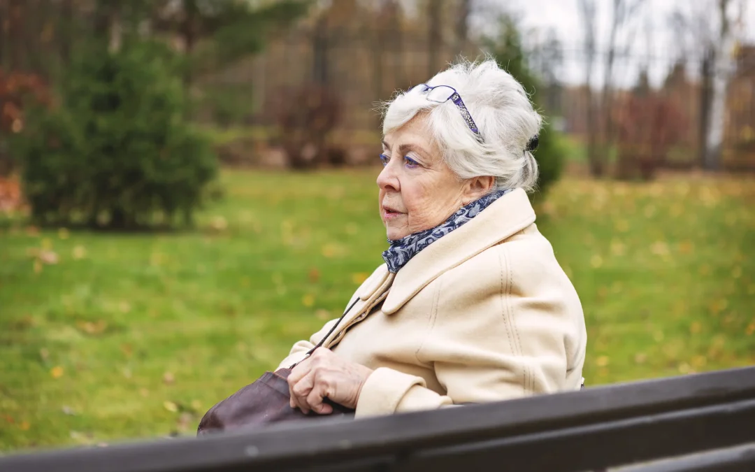 Elderly woman with white hair sits on a bench in a park, wearing a beige coat and scarf. She gazes thoughtfully ahead, surrounded by autumn trees.