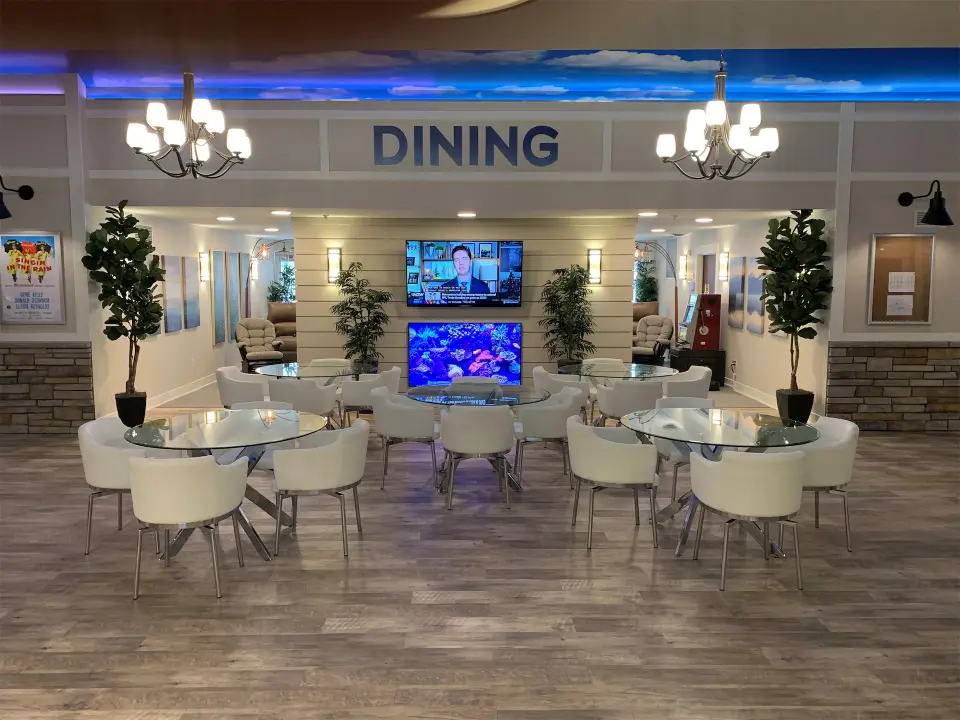 Modern dining area with glass tables and white chairs. Two TVs display news and aquarium visuals. Elegant chandeliers and potted plants enhance ambiance.